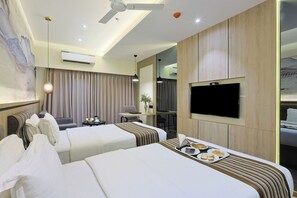 A hotel room with two beds, a flat-screen TV, a ceiling fan, an air conditioning unit, a desk with a chair, and a dining table with a tray of food. - Hotel President (Vadodara)