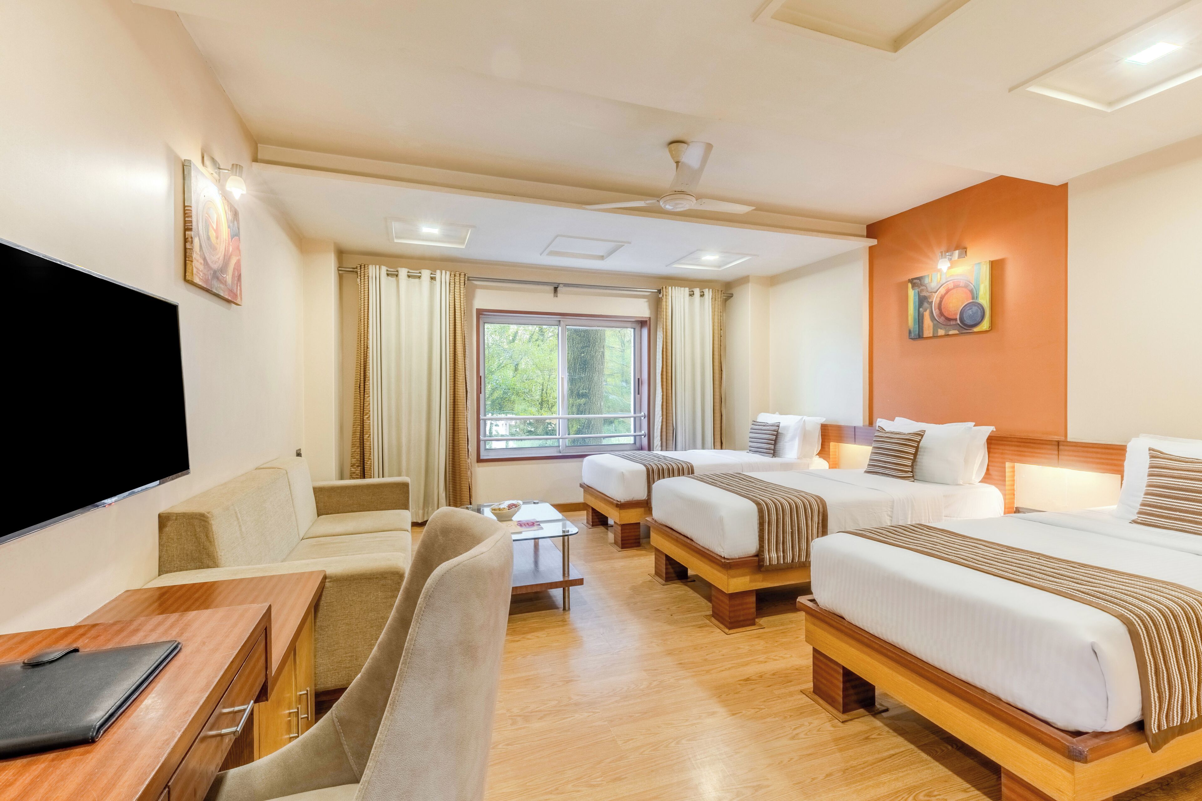 A hotel room with a flat-screen TV, a desk with a chair, two beds, a ceiling fan, and a window with curtains.