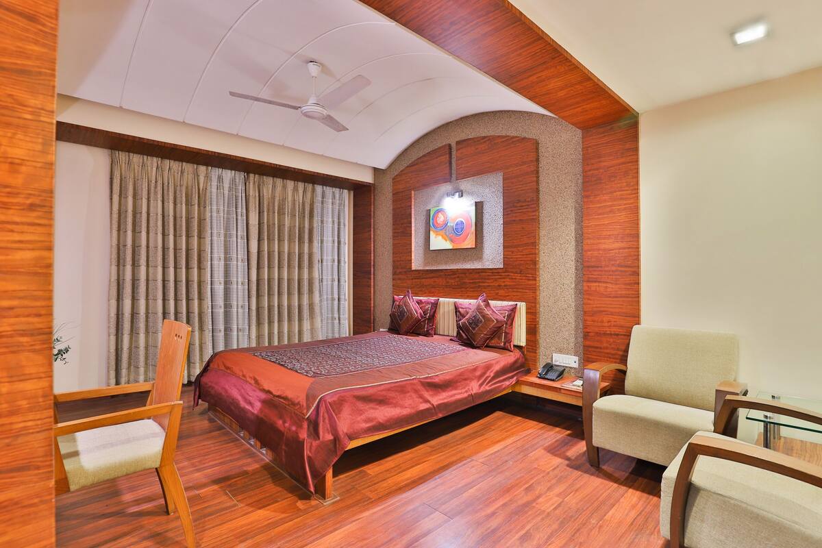 A hotel room with a wooden floor, a bed with a red bedspread, a ceiling fan, and a painting on the wall.