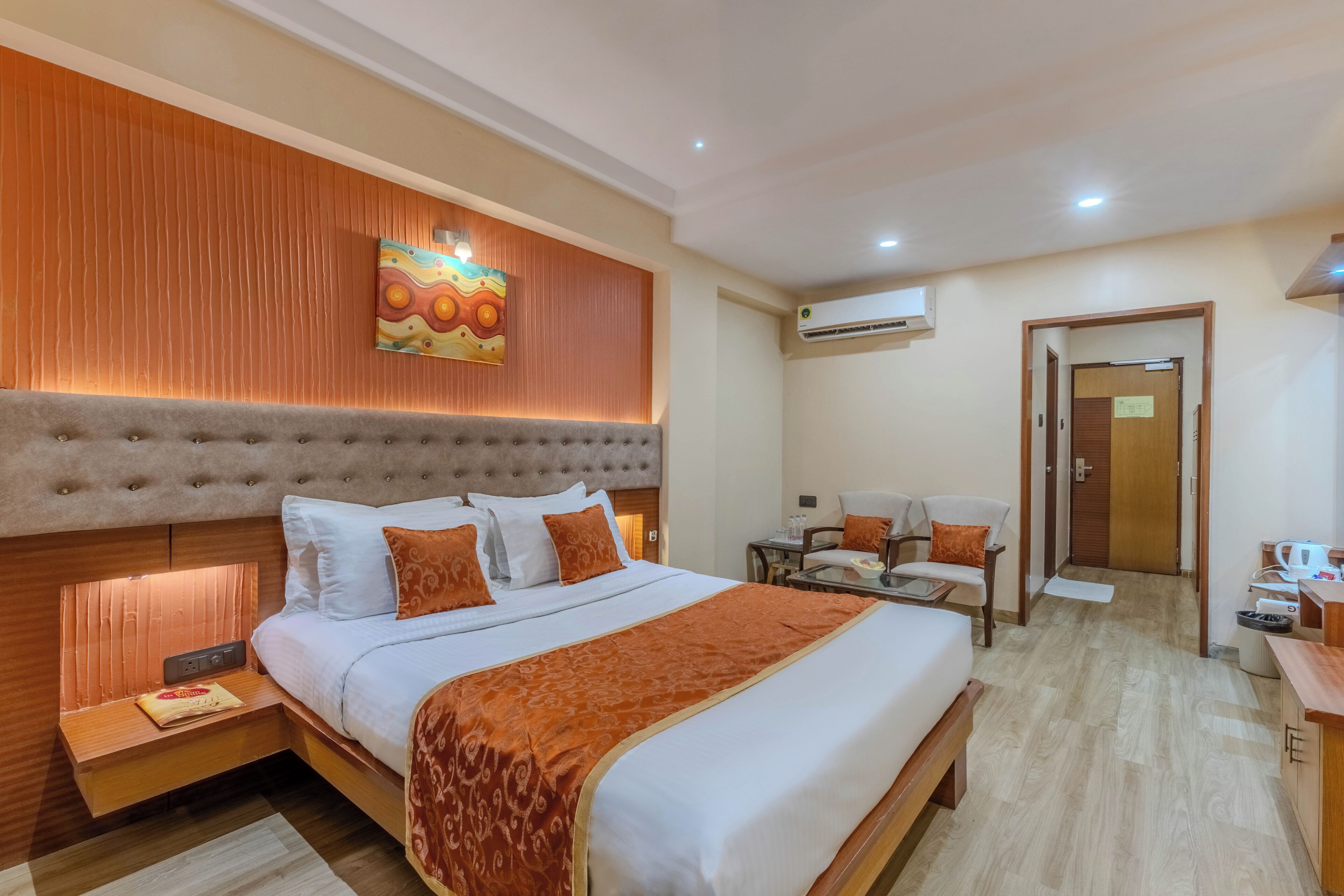 A hotel room with a large bed, a bedside table, a desk, a chair, and a small kitchenette.