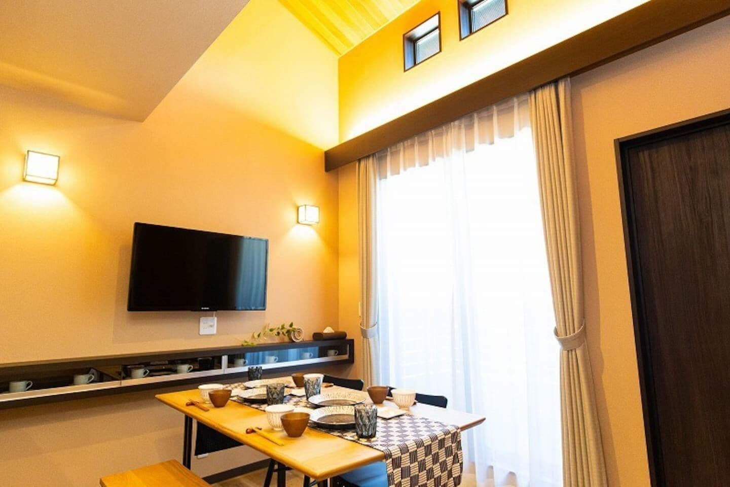 A hotel room with a dining table set for four, a flat-screen TV mounted on the wall, and a large window with curtains.