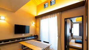 A hotel room with a wooden table, a flat-screen TV mounted on the wall, and a large window with curtains.