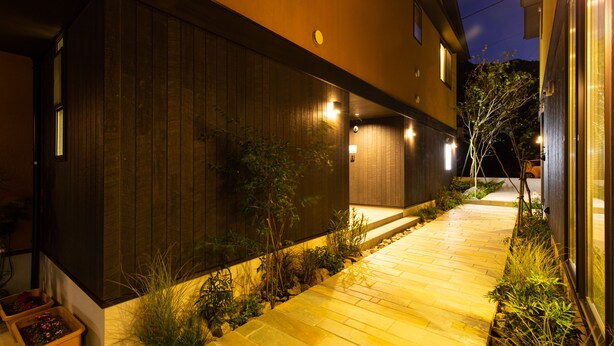 A nighttime view of a modern building with a wooden exterior, a paved pathway, and small plants along the sides.