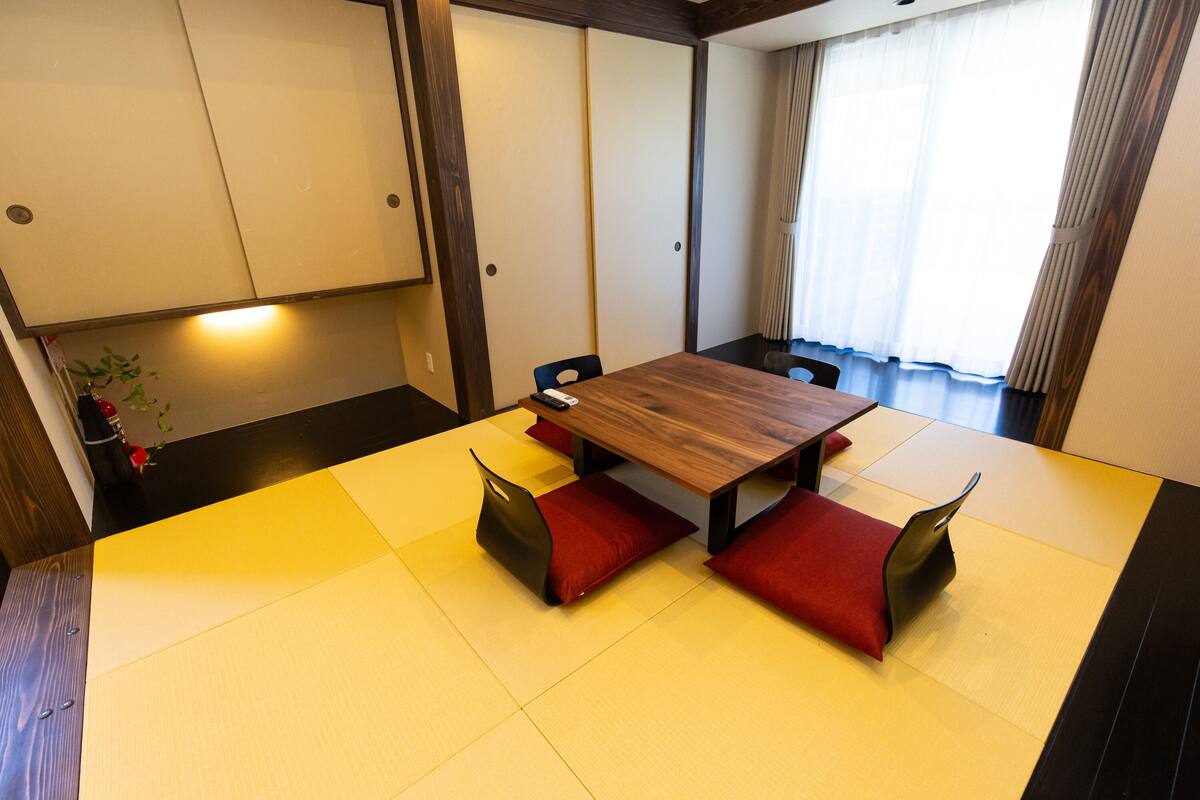 A traditional Japanese tatami room with a wooden table and red cushions, a sliding door, and a view of the outside through a window with curtains.