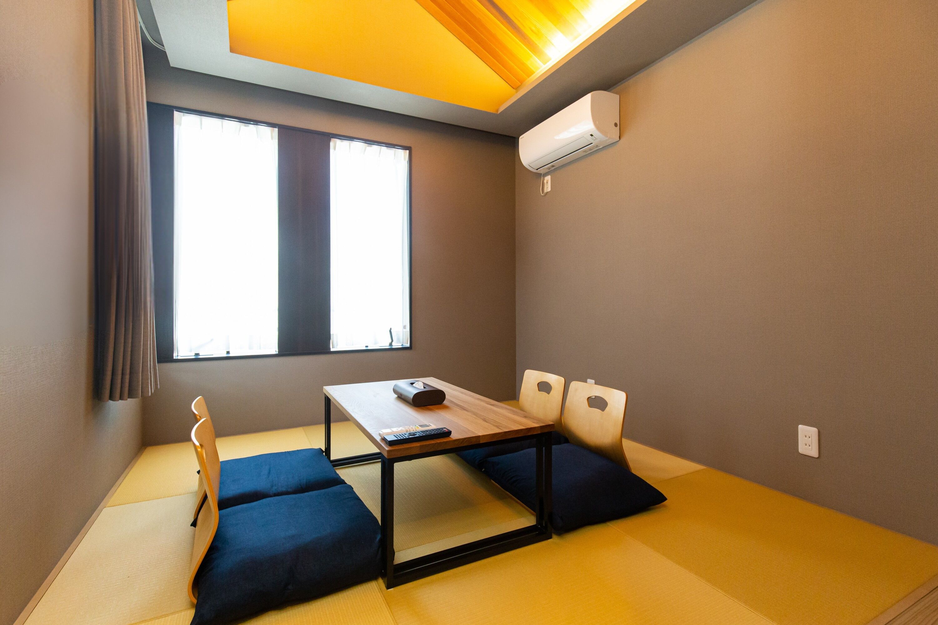 A room with tatami mat flooring, a low table with cushions, a window with curtains, and an air conditioning unit on the wall.