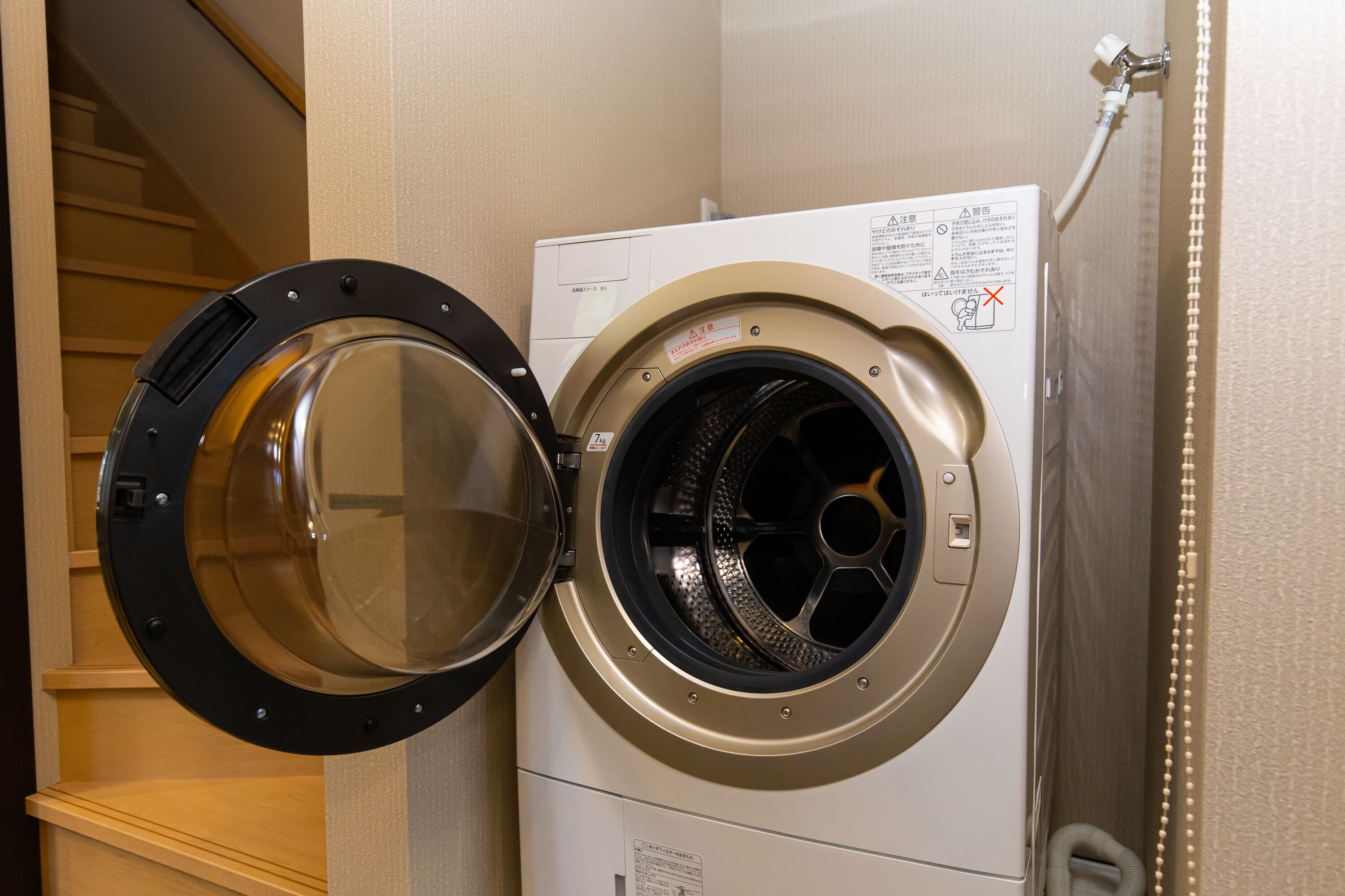 A front-loading washing machine in a laundry room with a wooden shelf and a staircase in the background.