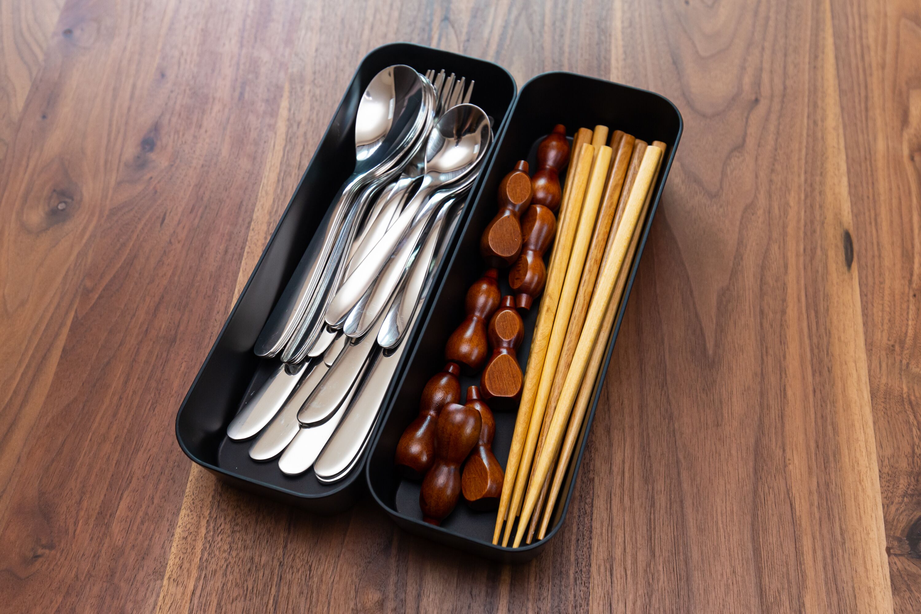 A black tray with silverware and wooden chopsticks on a wooden table.