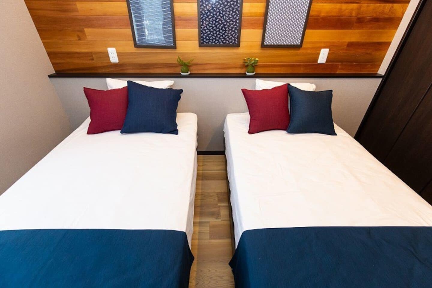Dos camas individuales con sábanas blancas y colchas azules, un cabecero de madera con paneles decorativos y pequeñas macetas con plantas en el cabecero.