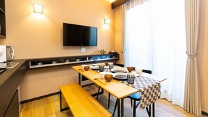 A compact hotel room with a kitchenette, a flat-screen TV mounted on the wall, a dining table set for two, and a large window with curtains.