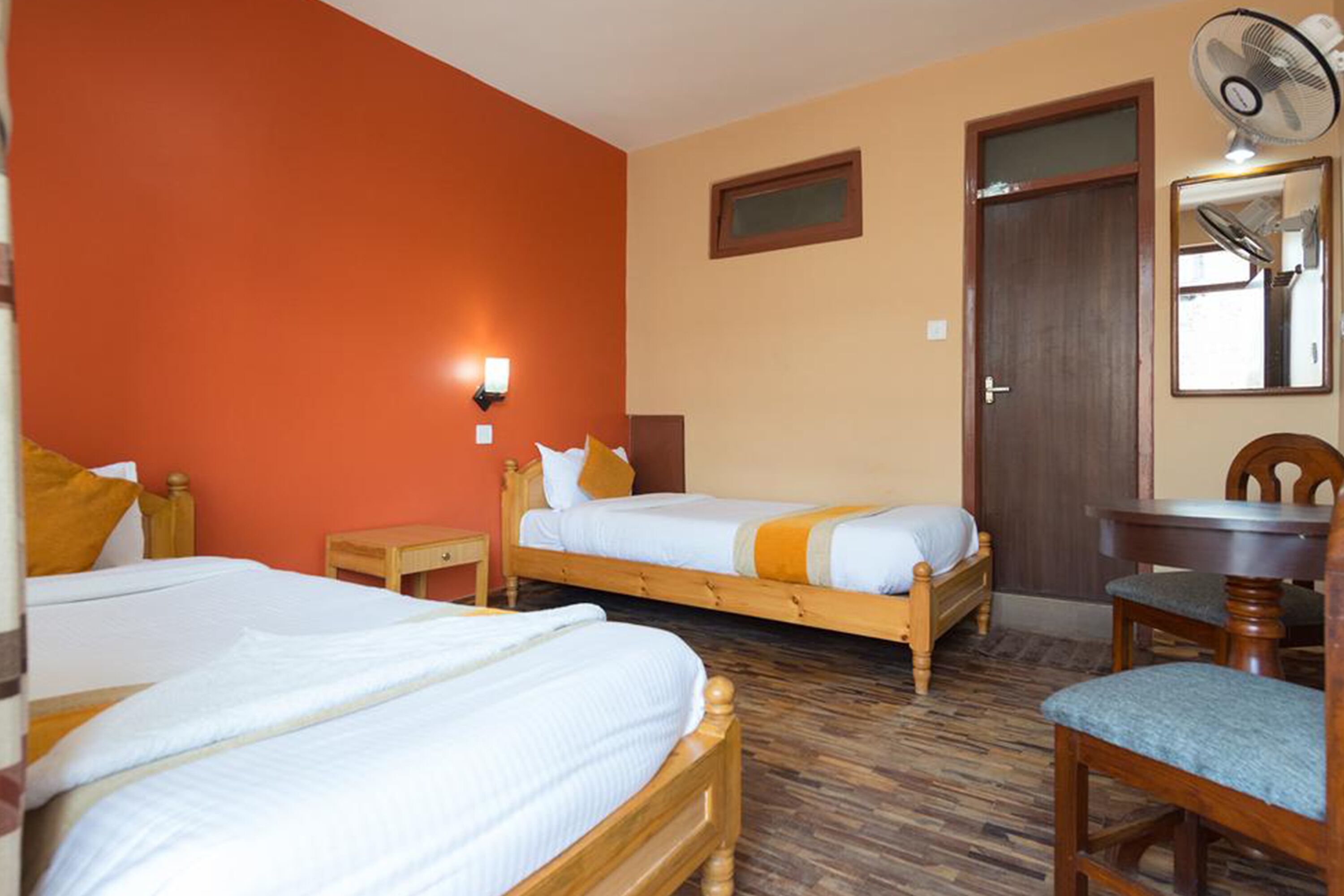Une chambre d’hôtel avec deux lits en bois, une chaise en bois, une petite table et un ventilateur fixé au mur.