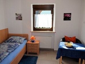 A small, well-lit room with a bed, a table with a basket, and a window with curtains.