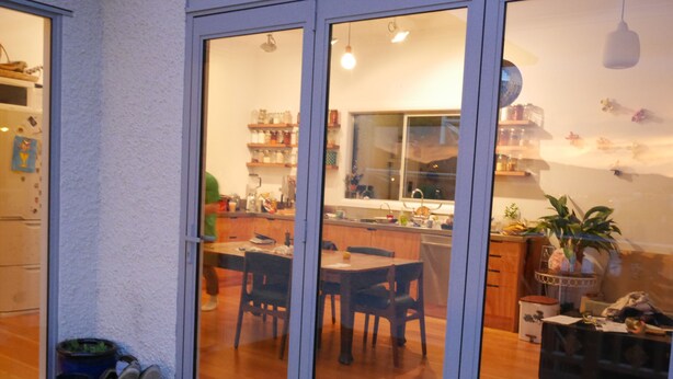 A kitchen with wooden cabinets, a dining table, and chairs. There is a sliding glass door leading to an outdoor area with a blue container and shoes. - ** Cake by the Ocean ** (St Clair)
