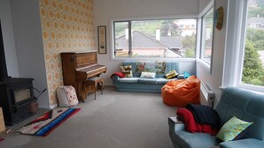 A living room with a piano, a blue sofa, a colorful rug, and a view of houses through the window. - ** Cake by the Ocean ** (St Clair)