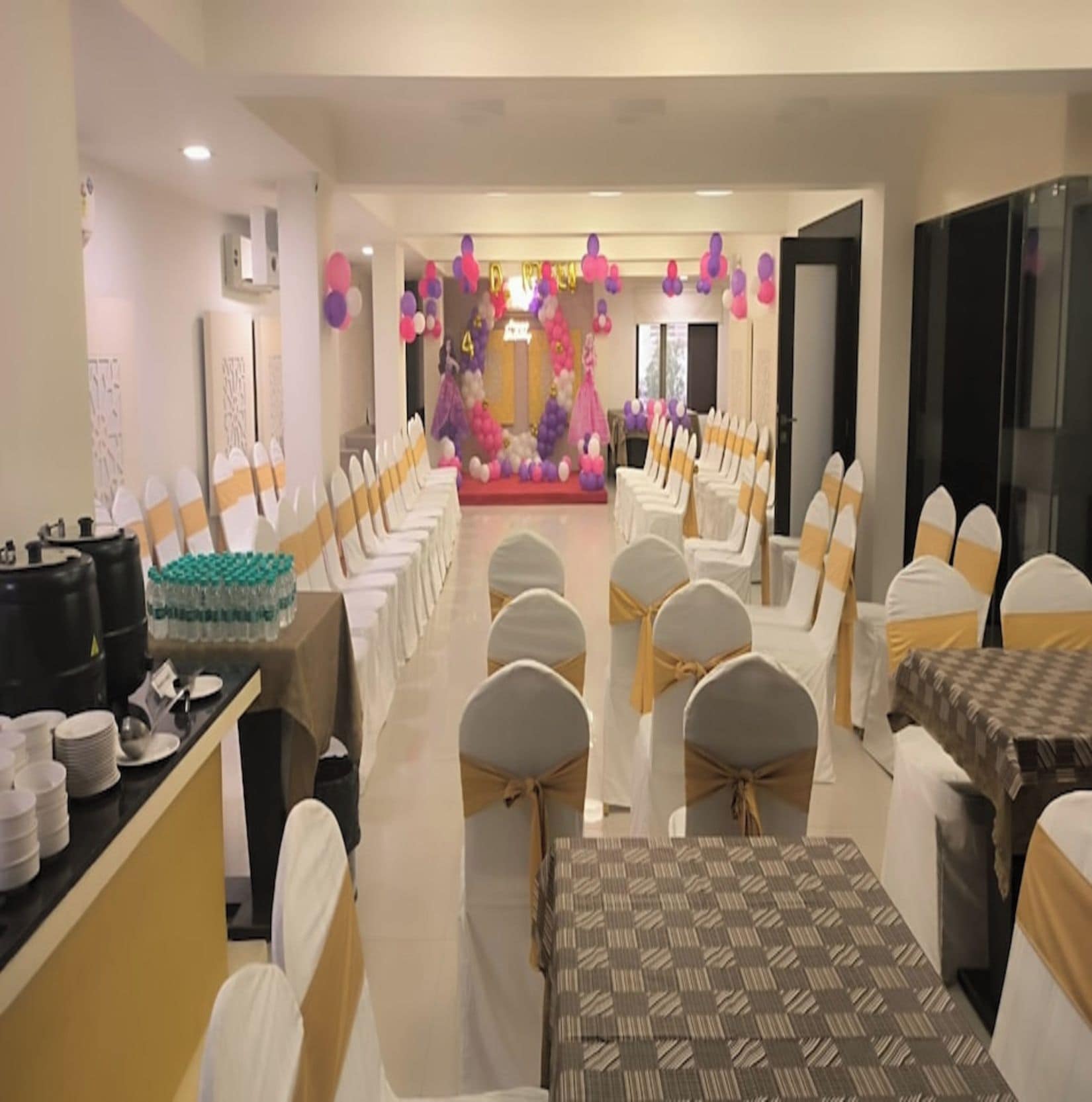 Una sala da banchetti con file di tavoli e sedie, un palco decorato con palloncini e un'area buffet.