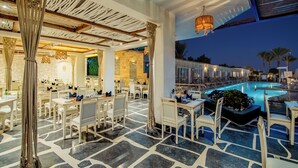 A restaurant with outdoor seating overlooking a pool, featuring stone flooring and white furniture.
