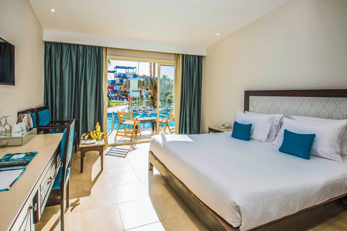 Une chambre d’hôtel avec un grand lit, un bureau avec une chaise, une petite table avec une chaise, et une vue sur l’espace piscine.