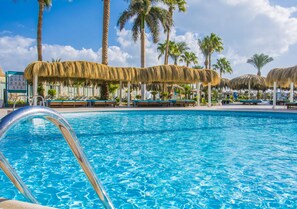 En swimmingpool med stråtag parasoller, palmer og liggestole.