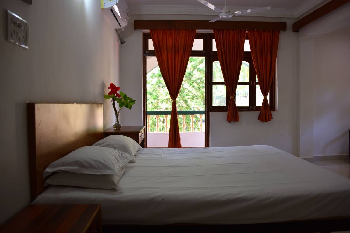 Quarto com cama, mesa de cabeceira, vaso com flores e janela com cortinas.