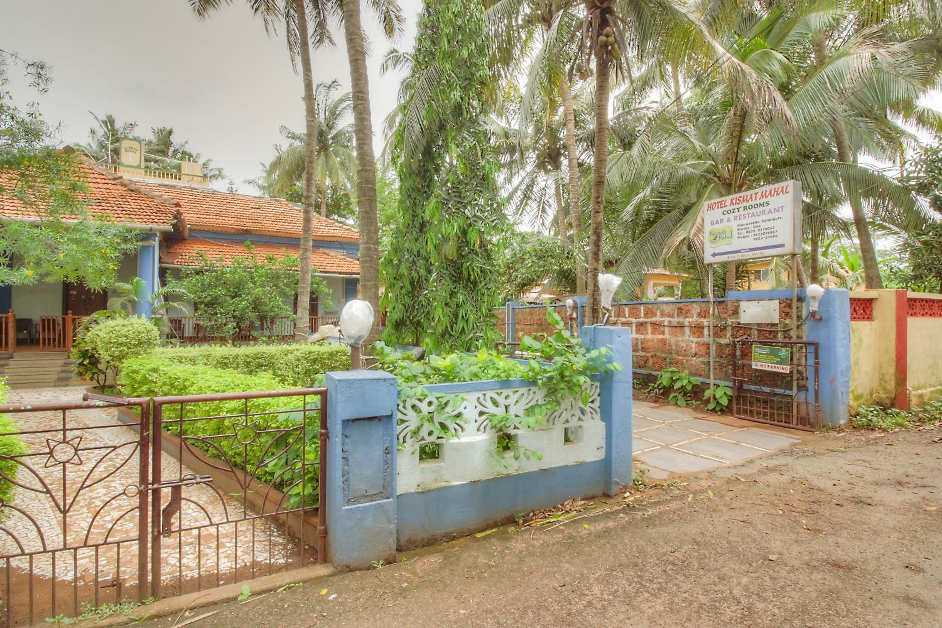 A hotel entrance with a signboard, a gated entrance, and a well-maintained garden with palm trees.