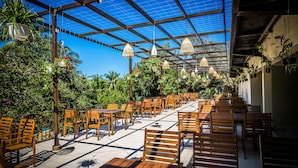 Patio esterno con tavoli e sedie di legno, immerso nel verde e decorato con piante pensili, sotto una struttura a copertura con vetro azzurro.