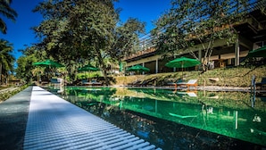 Area piscina con superficie d'acqua limpida, circondata da vegetazione rigogliosa e ombreggiata da grandi ombrelloni verdi.