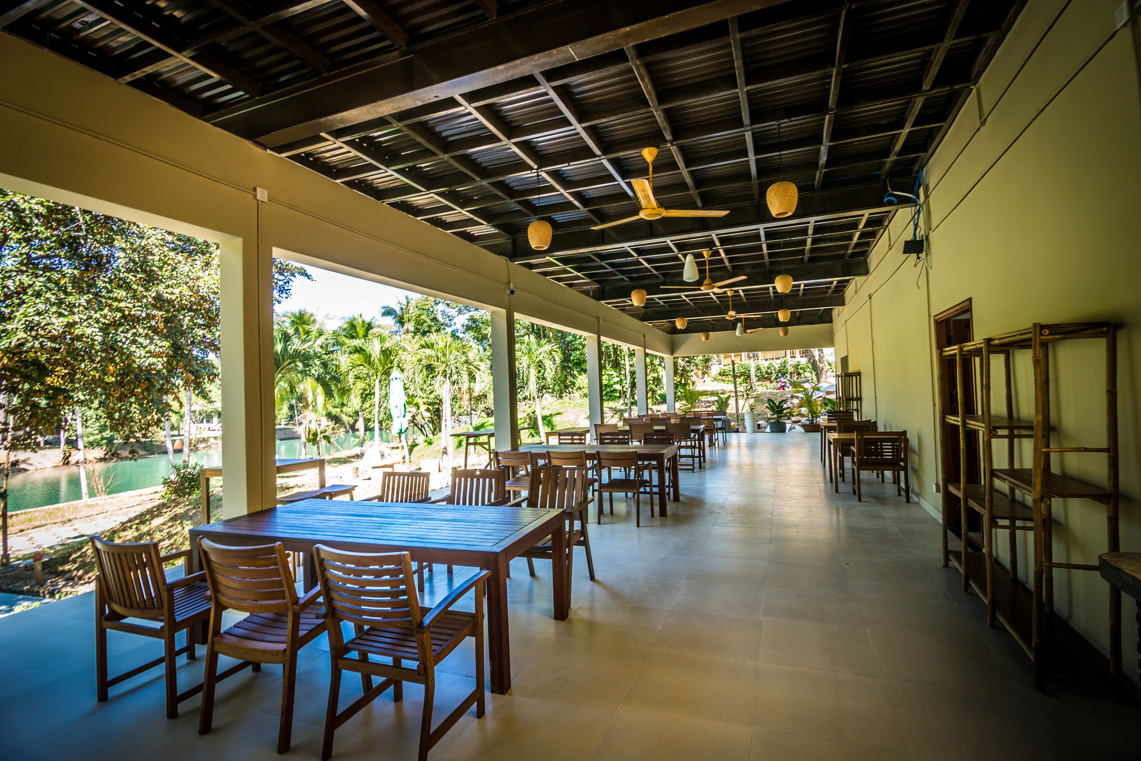 A spacious covered dining area with wooden tables and chairs, large windows, and a view of a lake and greenery.