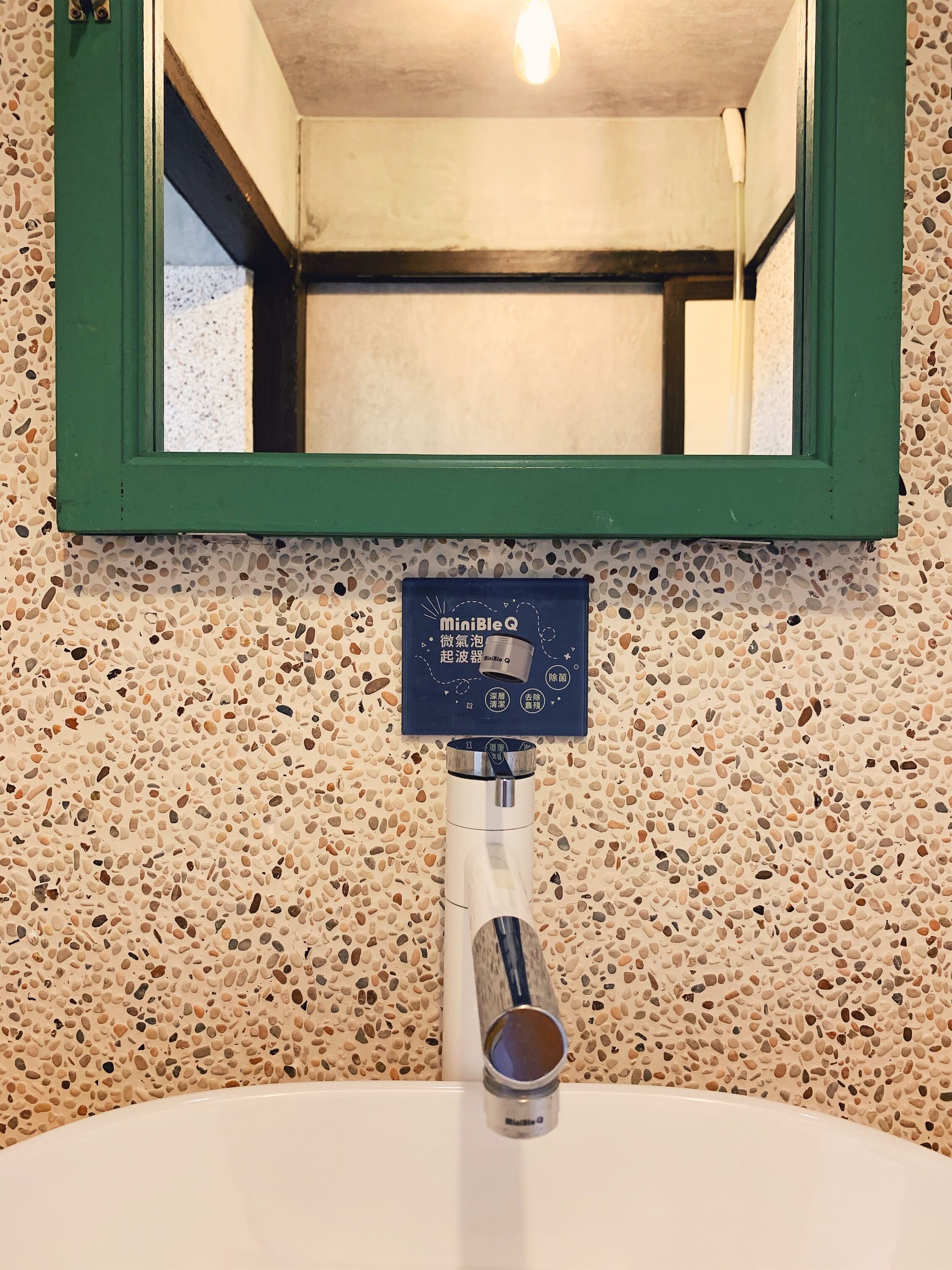 Un baño con pared de azulejos veteados, espejo con marco verde y lavabo blanco con sistema de filtración de agua MiniBleQ.