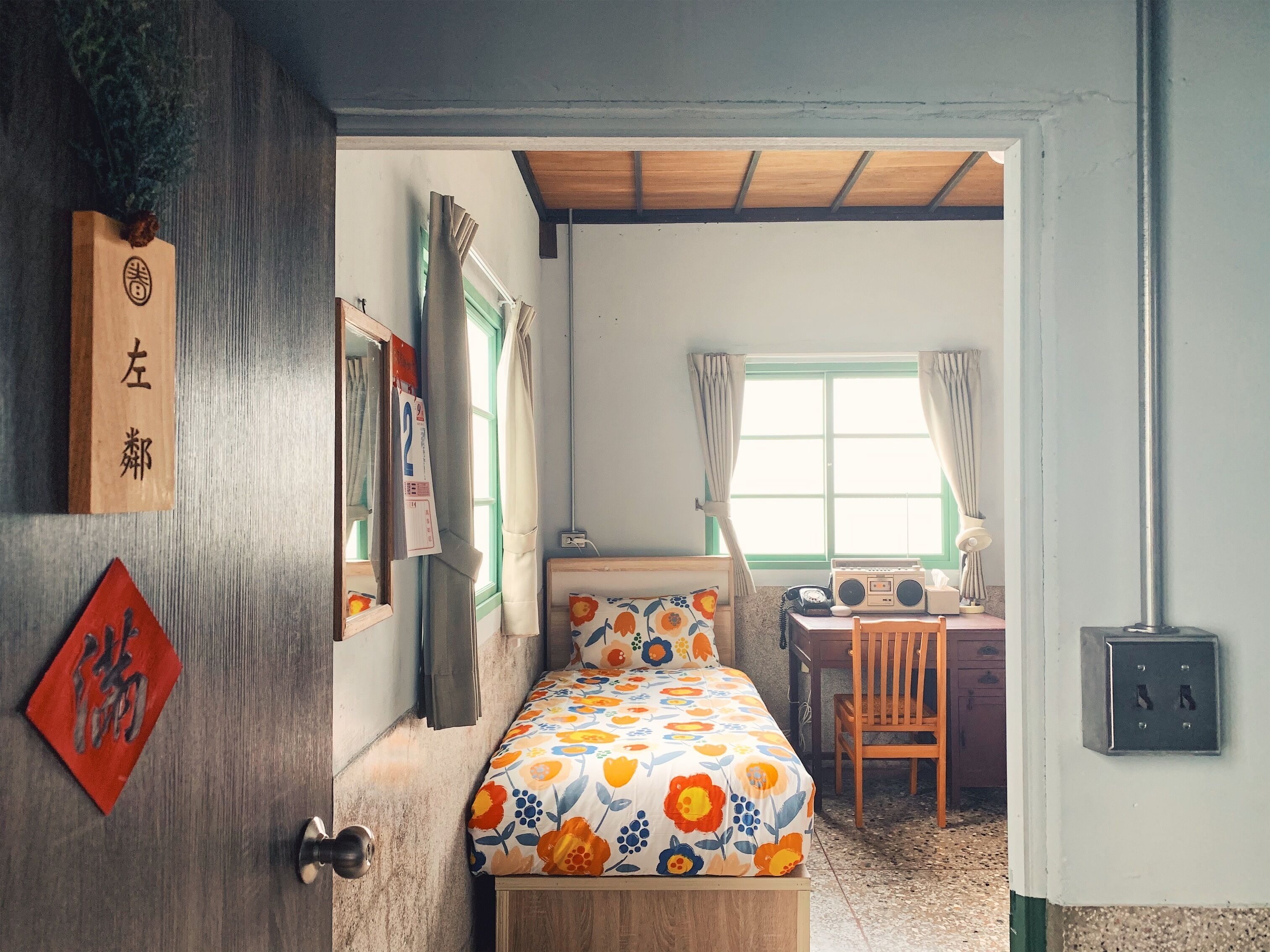 Une pièce comprenant un lit, un bureau et une chaise. Il y a une fenêtre avec des rideaux, une porte avec une poignée et un mur avec un interrupteur.