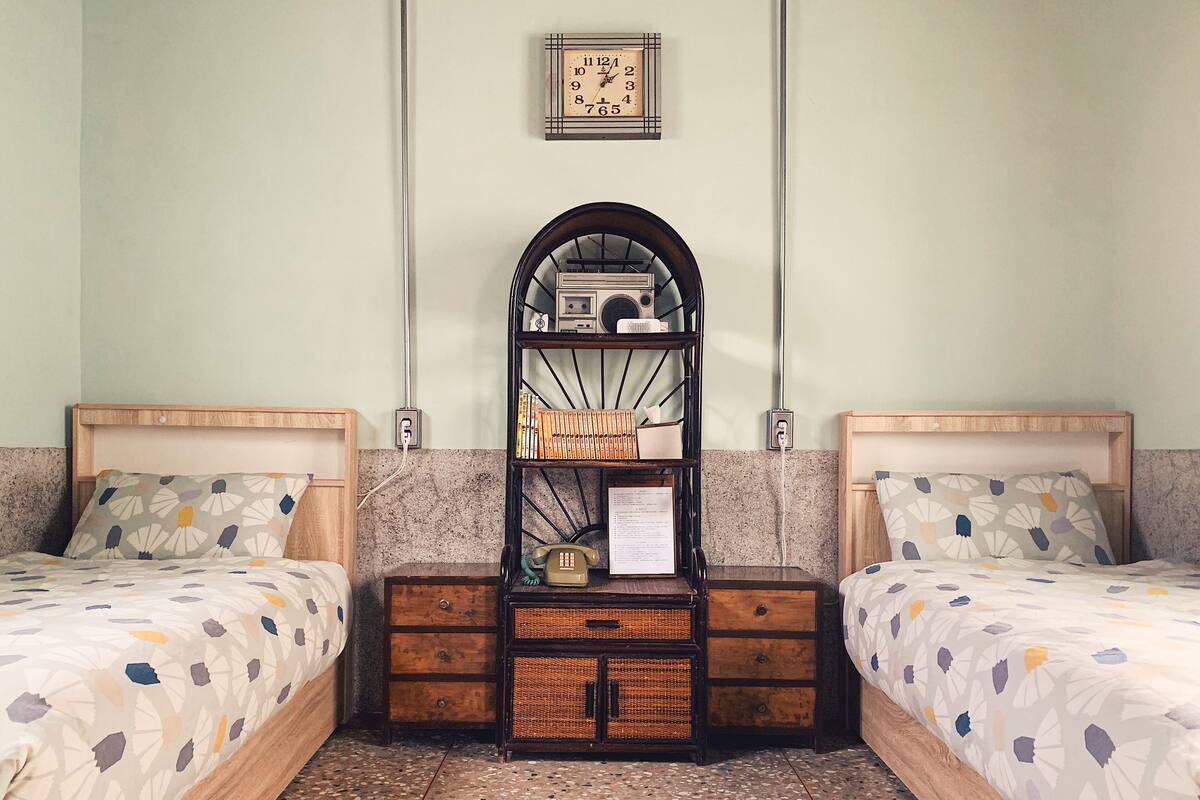 Een kamer met twee bedden, een wandklok, een plank met boeken en een telefoon, en een betegelde vloer.