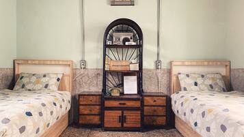 A room with two beds, a wall clock, a shelf with books and a telephone, and a tiled floor.