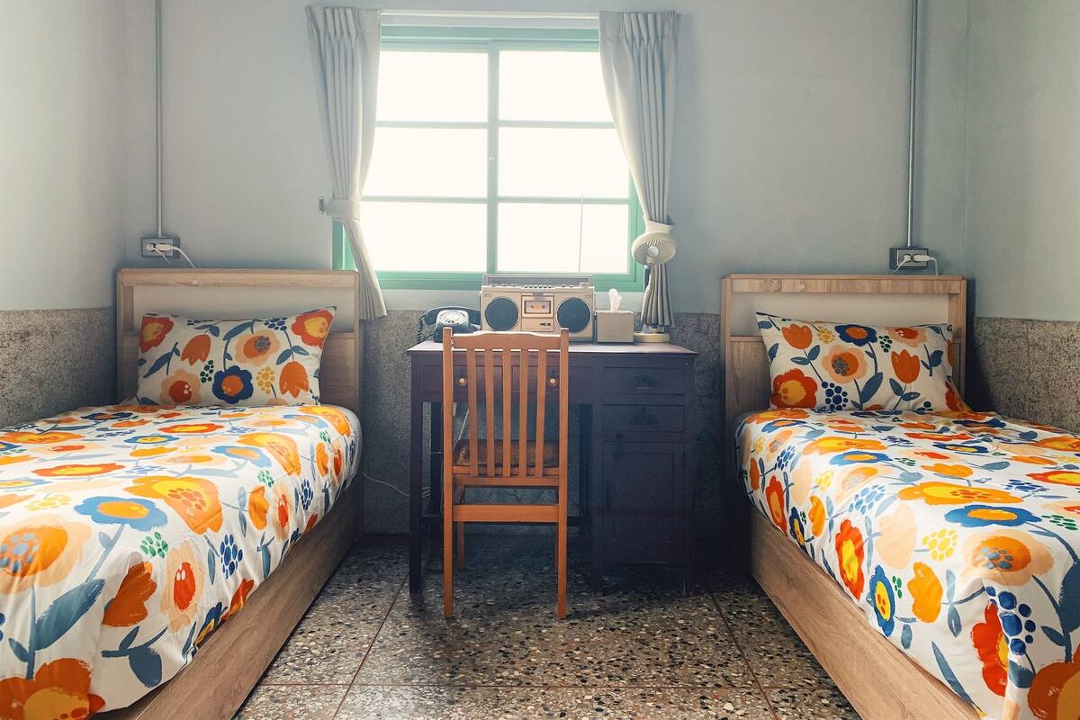Een kamer met twee bedden, een bureau, een stoel, een radio en een raam met gordijnen.