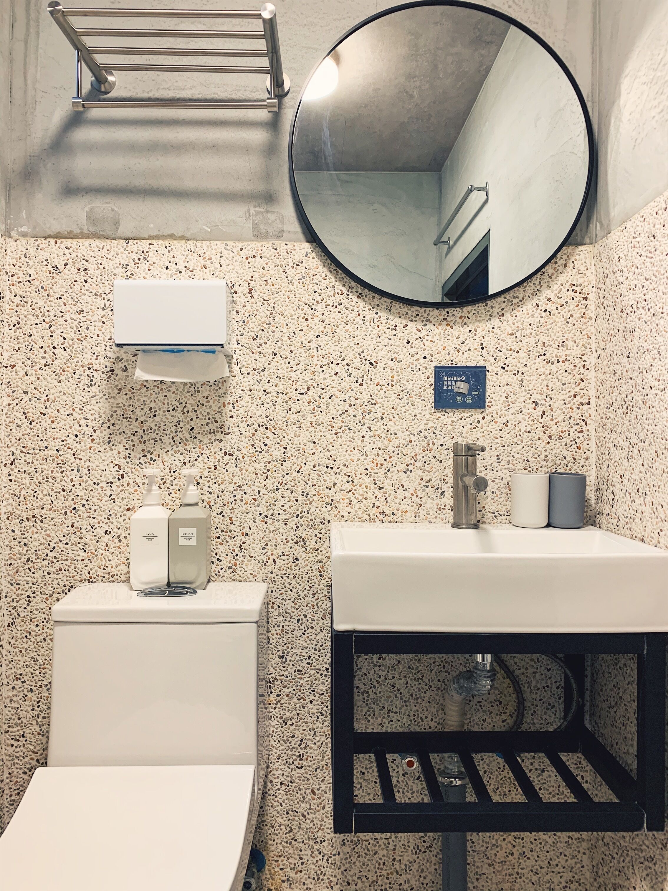Ein Badezimmer mit Toilette, Waschbecken und Spiegel. Über der Toilette befindet sich ein Handtuchhalter und am Waschbecken ein Seifenspender.