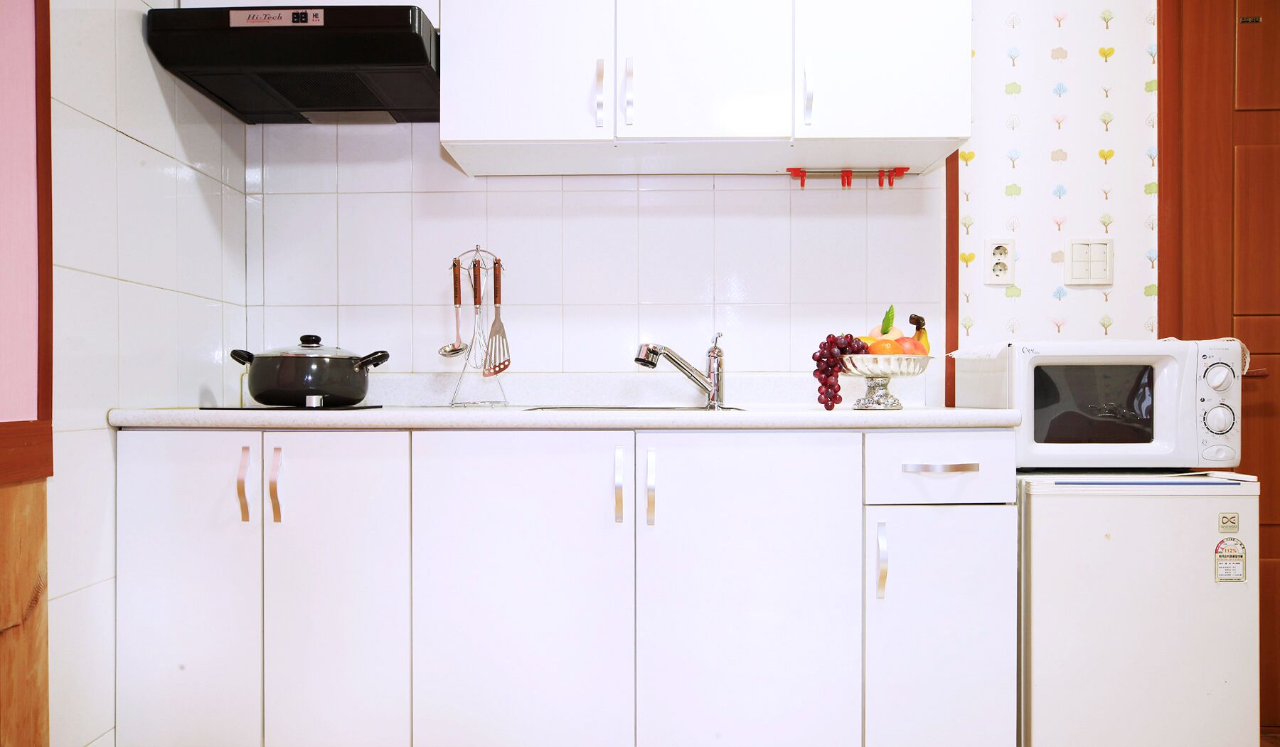 Une cuisine avec des meubles blancs, une cuisinière noire, un évier avec un robinet, un panier de fruits et un four à micro-ondes.