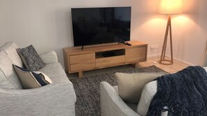 A modern living room with a flat-screen TV on a wooden TV stand, a grey sofa with cushions, a floor lamp, and a rug. - Baden 70 - Rainbow Shores. Ground Floor. Air Con. Overlooking lap pool. Wi-Fi (Rainbow Beach)