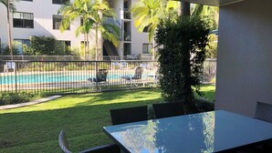 A view of a pool area with lounge chairs, a fenced pool, and a building with multiple balconies in the background. - Baden 70 - Rainbow Shores. Ground Floor. Air Con. Overlooking lap pool. Wi-Fi (Rainbow Beach)