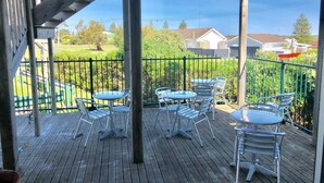A wooden deck with metal chairs and tables, a metal railing, and a view of a green landscape and houses in the background. - Pinehaven Motel (Nelson)