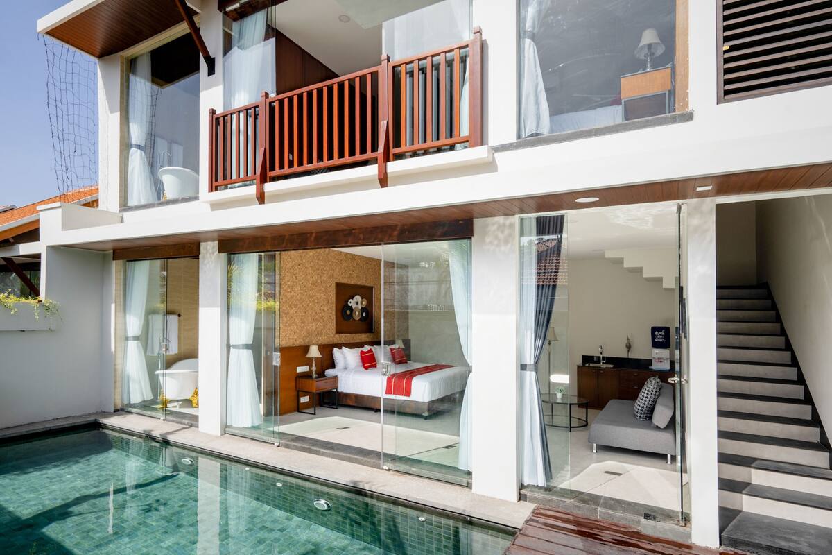 A modern two-story house with a glass-enclosed bedroom, a balcony with wooden railings, and a swimming pool.