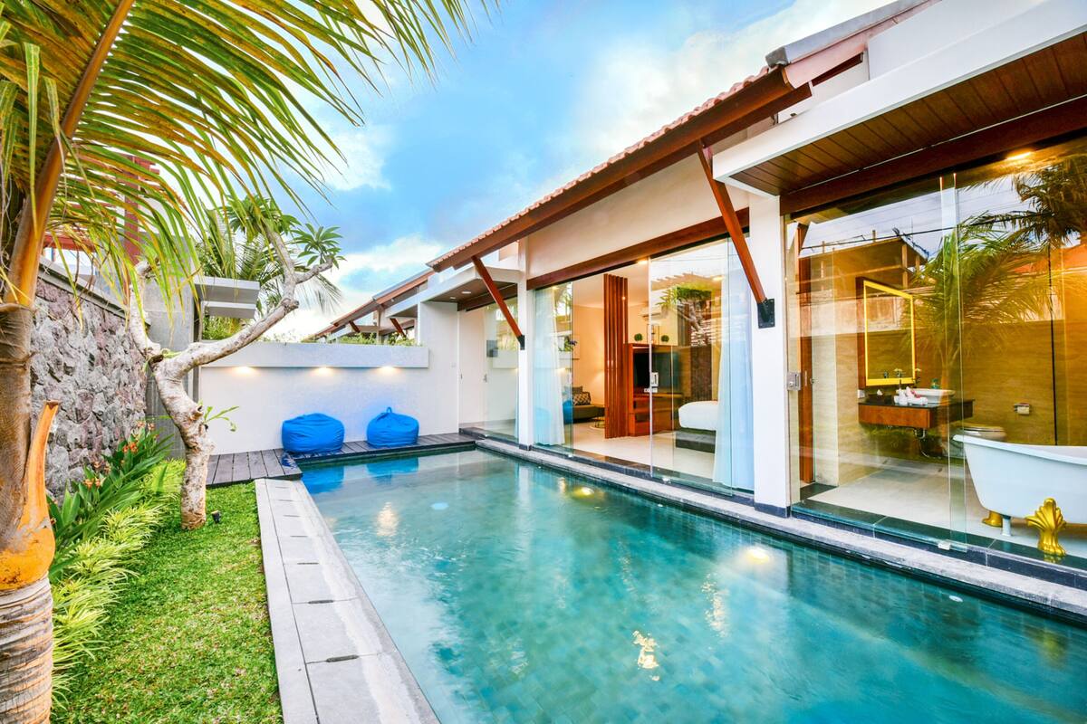 A modern pool area with clear blue water, a wooden deck, and a glass-enclosed bathroom with a golden bathtub.
