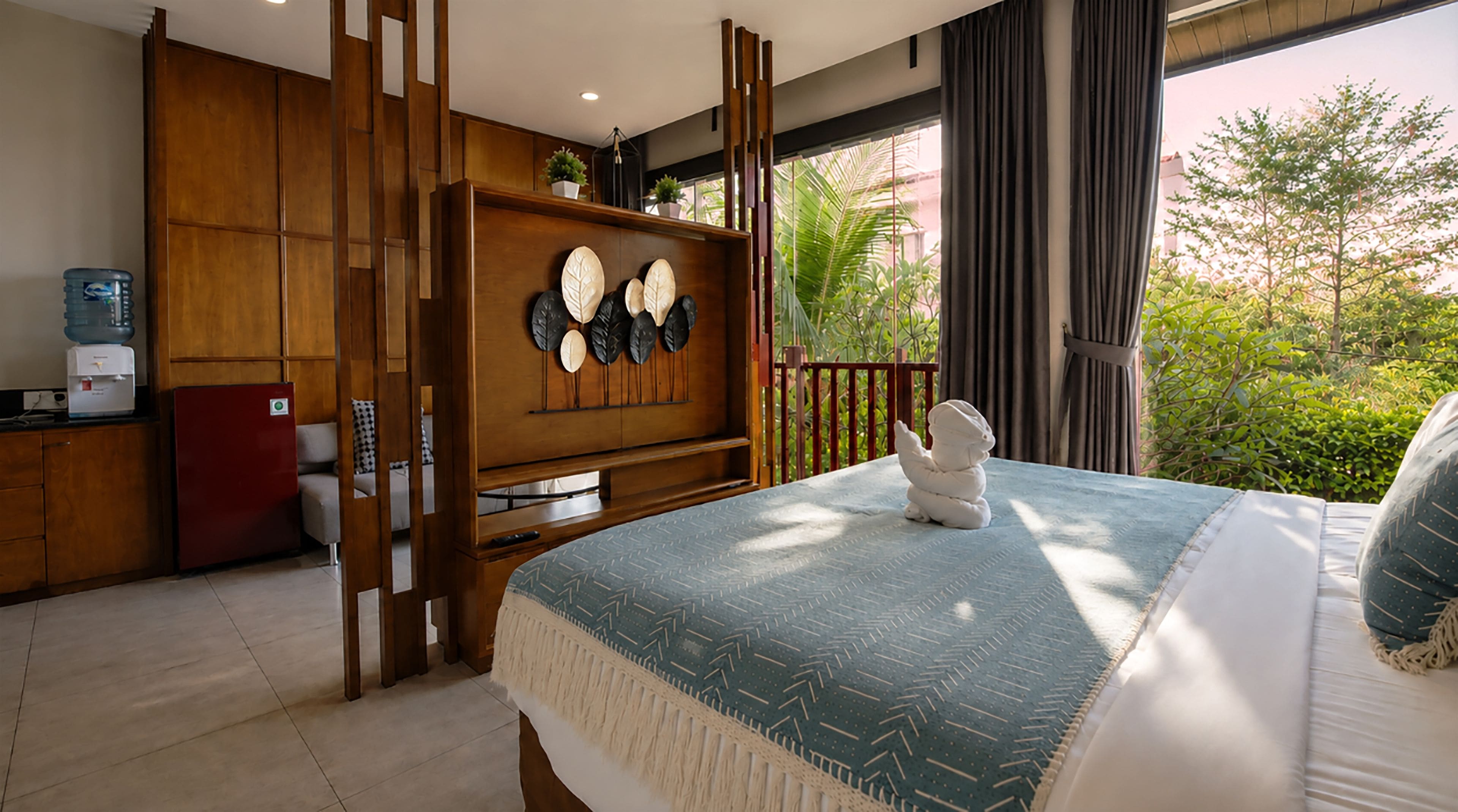 A bedroom with a bed, a wooden cabinet, a water dispenser, and a view of greenery outside.