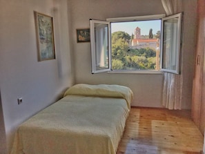 A small room with a single bed, a window with curtains, and a view of a building and trees outside. - Vila Keti is set in Drvenik Veliki. (Drvenik Veli)