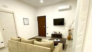 A living room with a beige sofa, a coffee table, a TV mounted on the wall, and a door in the background. - Villa Elisa Apartments (Minturno)