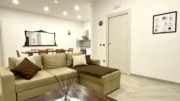 A modern living room with a sofa, a coffee table, and a dining area in the background. - Villa Elisa Apartments (Minturno)