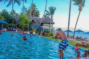A swimming pool area with people swimming and lounging, surrounded by palm trees and a building with a thatched roof.