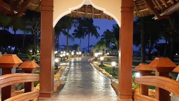 A pathway with a thatched roof structure, palm trees, and illuminated lights.