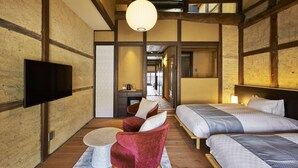 Une chambre d’hôtel avec un plancher en bois, un téléviseur fixé au mur, une chaise rouge, deux lits avec une literie à motifs et un luminaire suspendu.