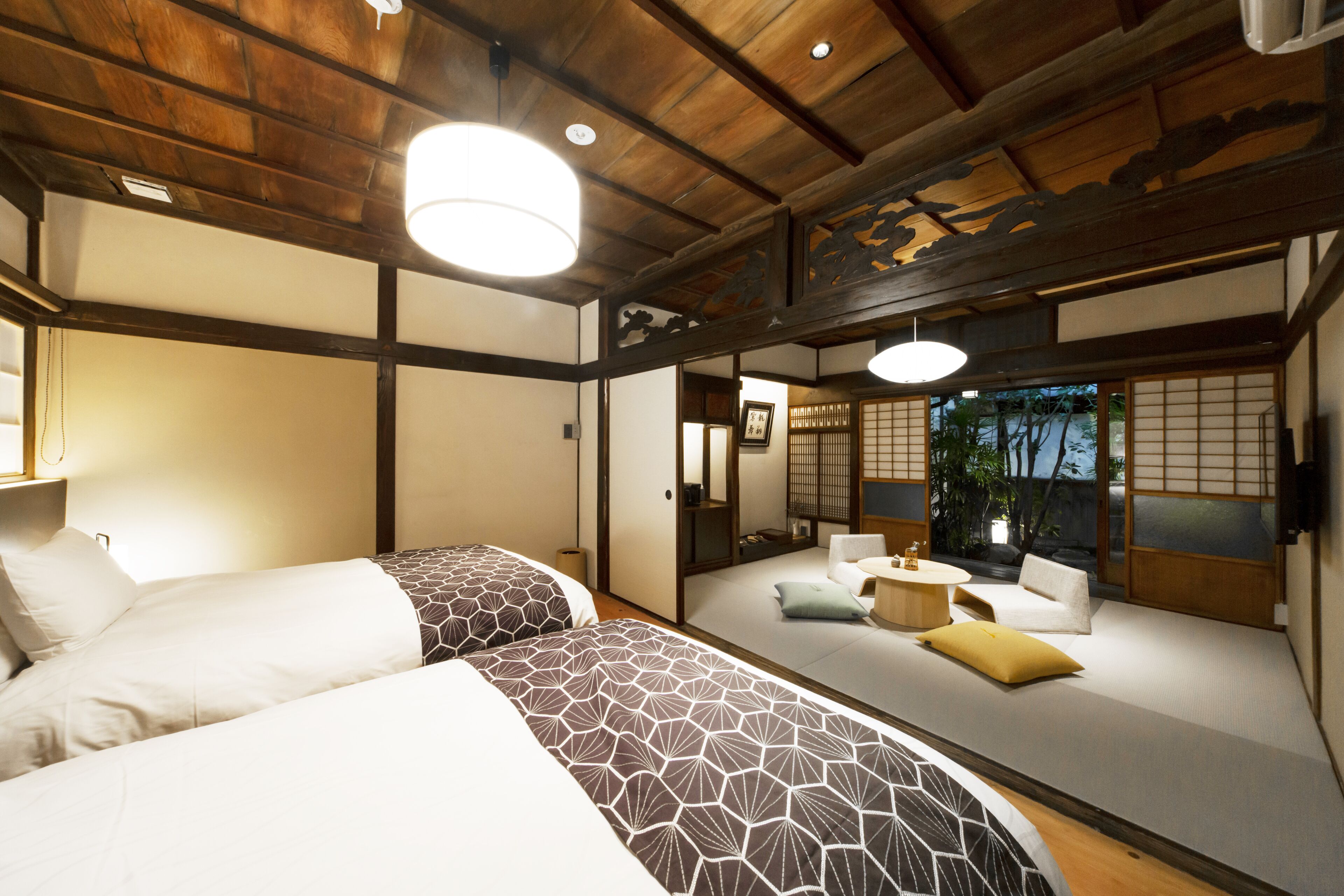 Ein traditioneller japanischer Raum mit Tatami-Bodenbelag, niedriger Holzdecke und Schiebetüren. Zwei Betten mit gemusterten Bettwäsche sowie eine kleine Sitzecke mit Kissen sind vorhanden.