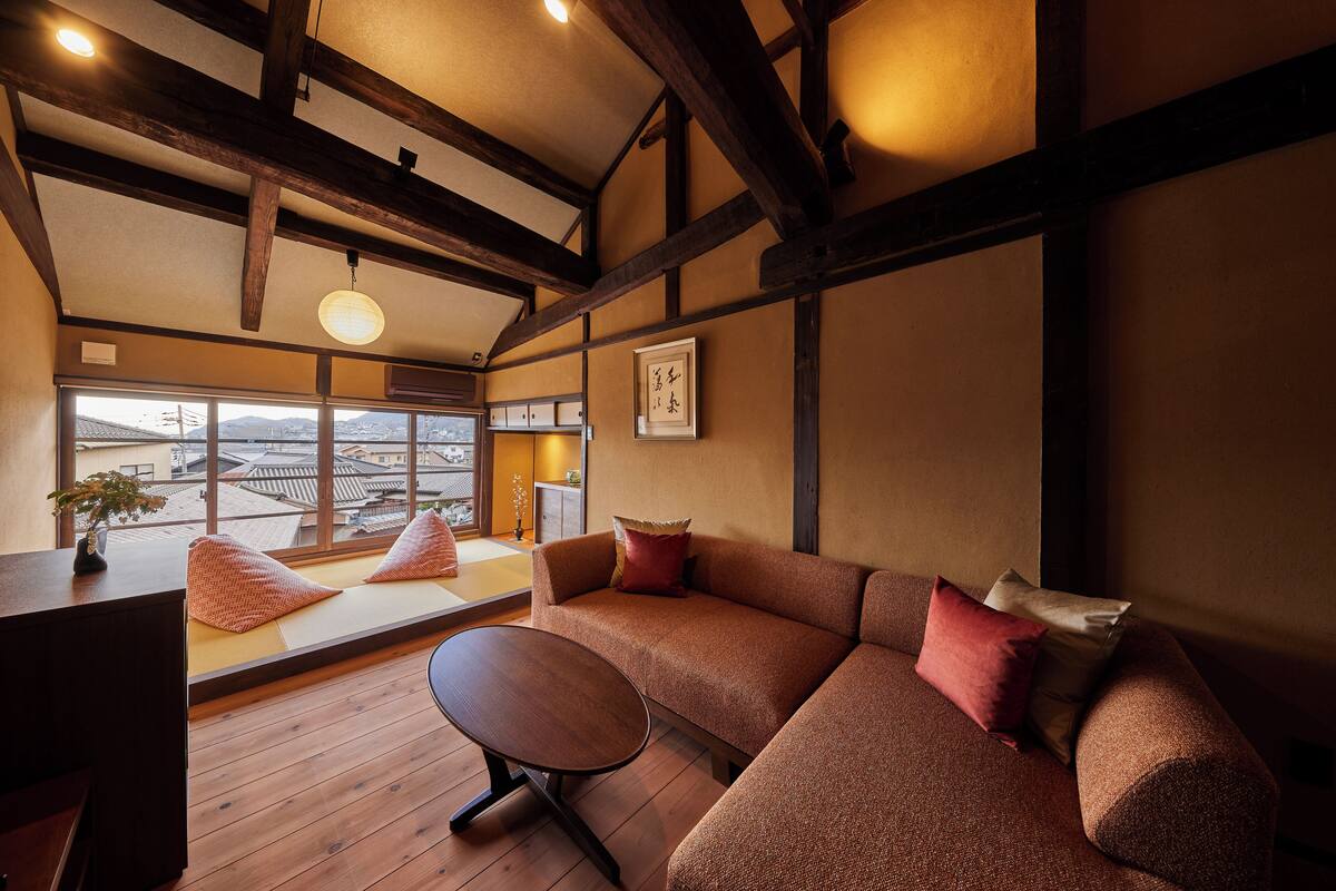 A traditional Japanese room with tatami flooring, a low wooden table, a sofa with cushions, and a large window offering a view of the city.