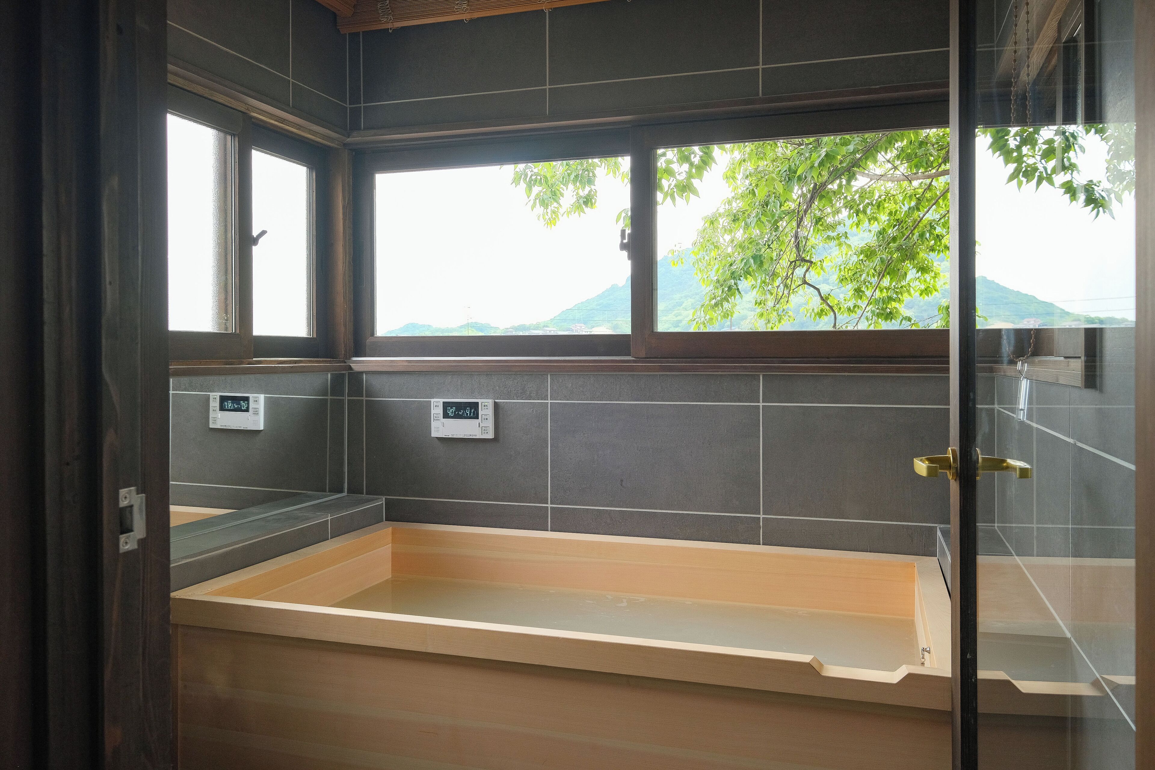 A bathroom with a bathtub, two digital thermostats, and a view of greenery outside.