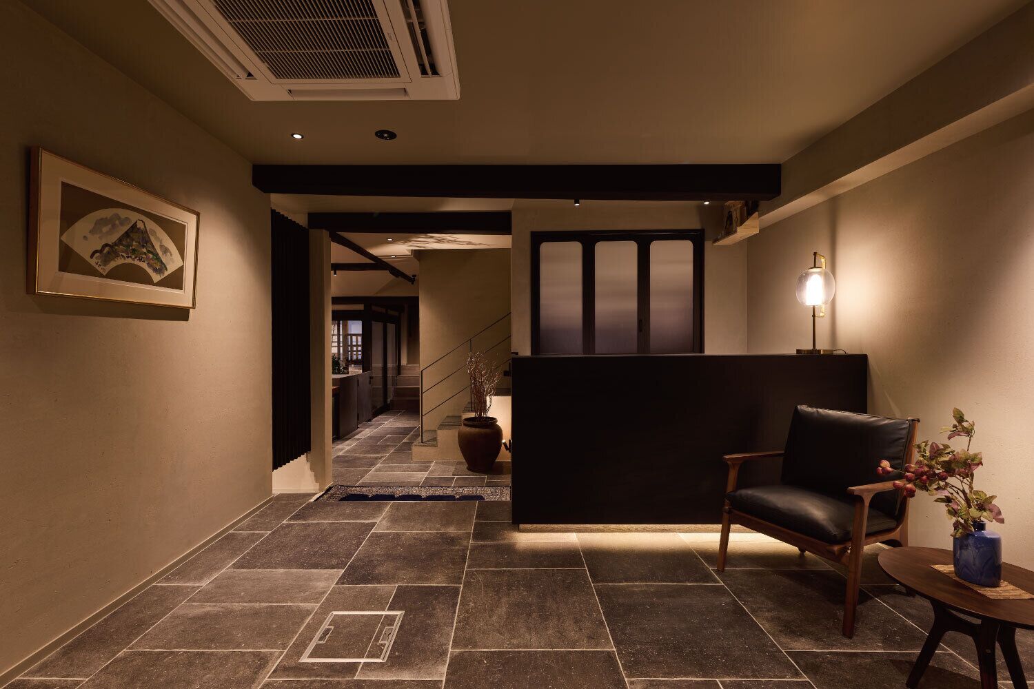 A hallway with a staircase, a reception desk, a chair, and a table with a vase.