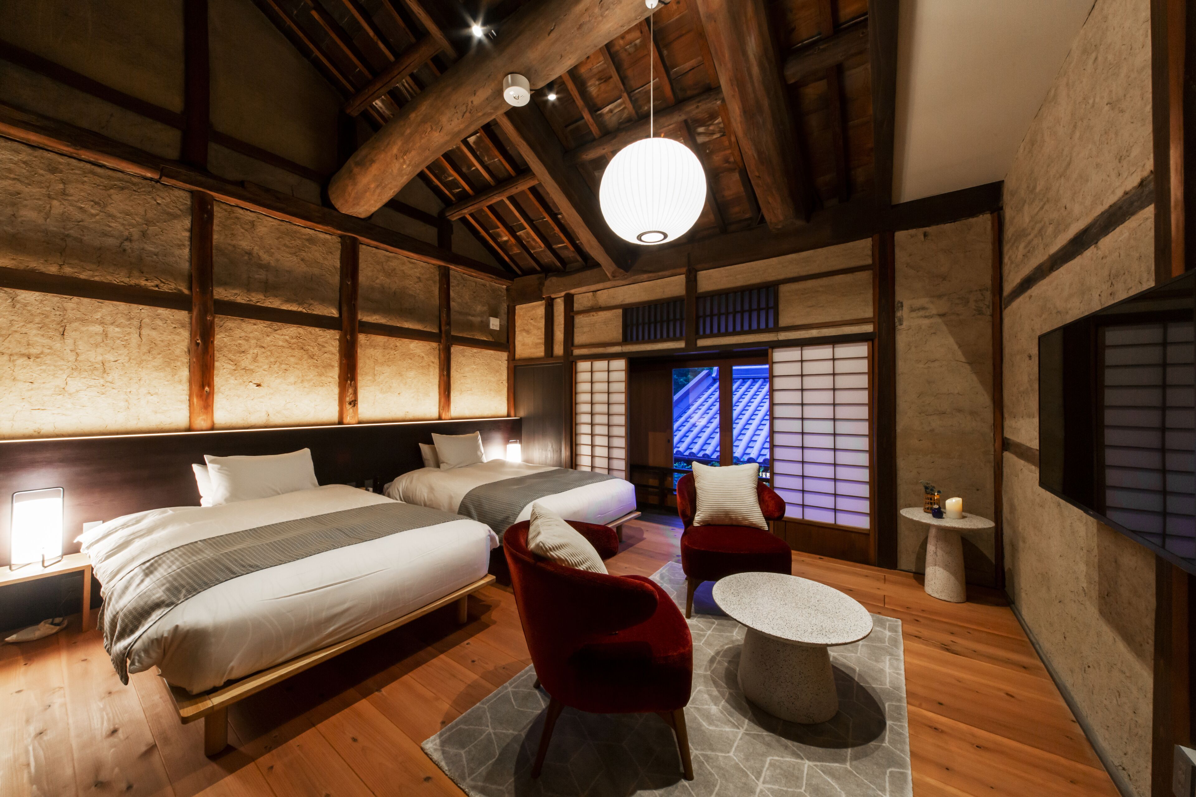 A traditional Japanese-style room with two beds, a red chair, a round table, and a wooden ceiling.